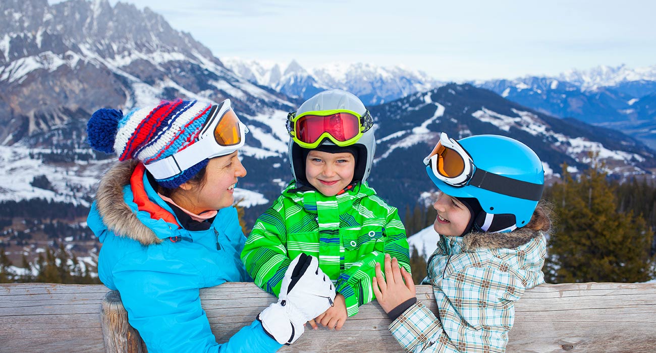 Mutter und zwei Kinder in Skiausrüstung mit Helm lehnen sich an einen Holzzaun